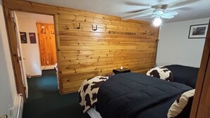 A room with a wooden wall, a bed with a cow print blanket, a ceiling fan, and a door leading to another room. - Majestic Recreational Lodging w/ Snowmobile and Side x Side rentals on Long Lake (Sinclair)