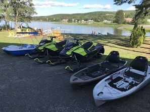 Une rangée de motoneiges et un petit bateau sont stationnés sur une zone de gravier, près d’un lac.