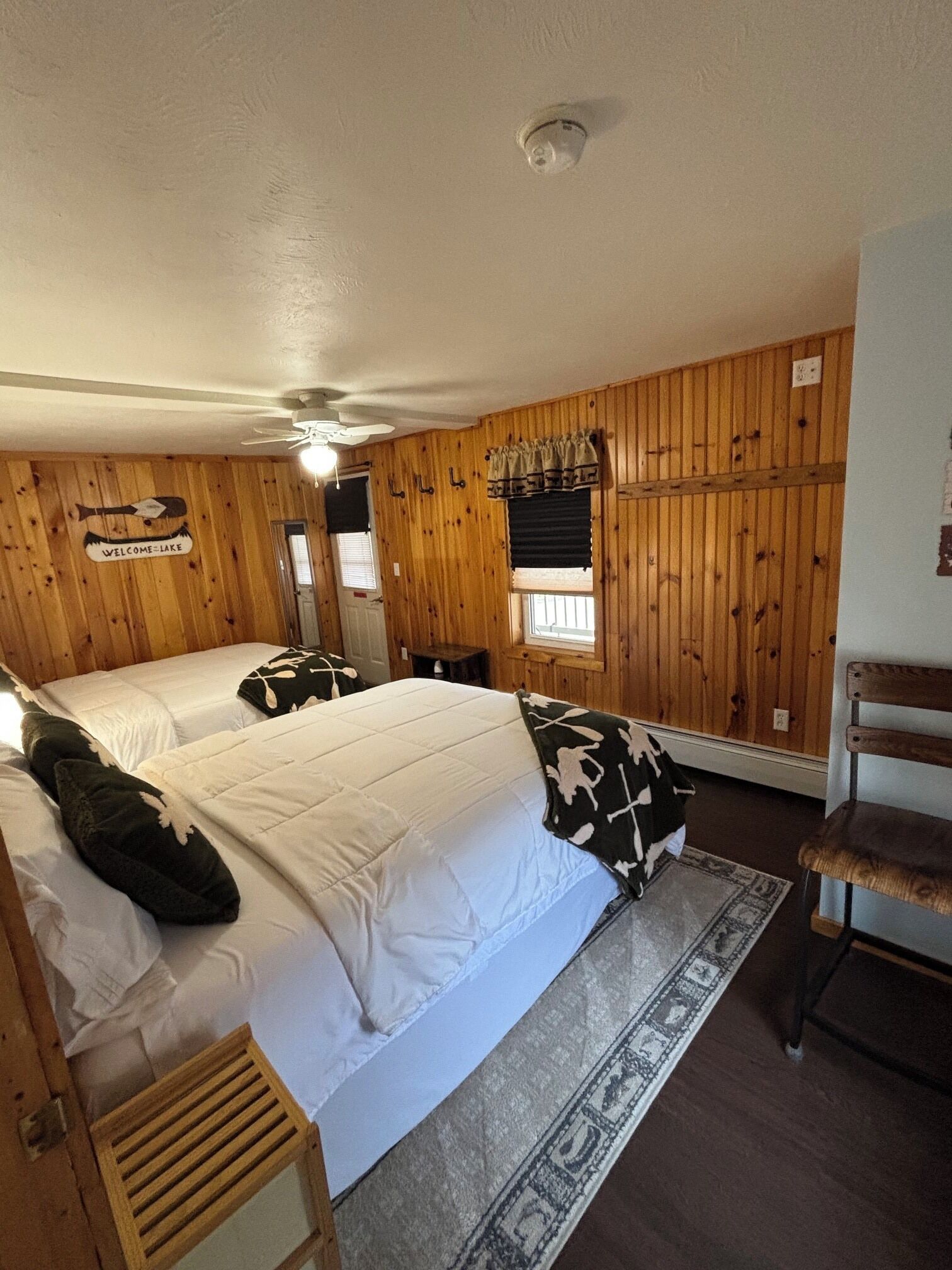 A room with wooden paneling, two beds with white bedding, a ceiling fan, a window with blinds, and a small table.