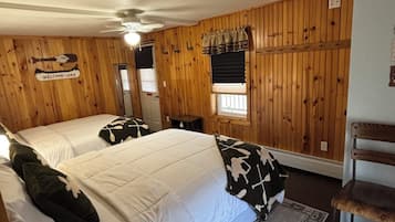 A room with wooden paneling, two beds with white bedding, a ceiling fan, a window with blinds, and a small table.