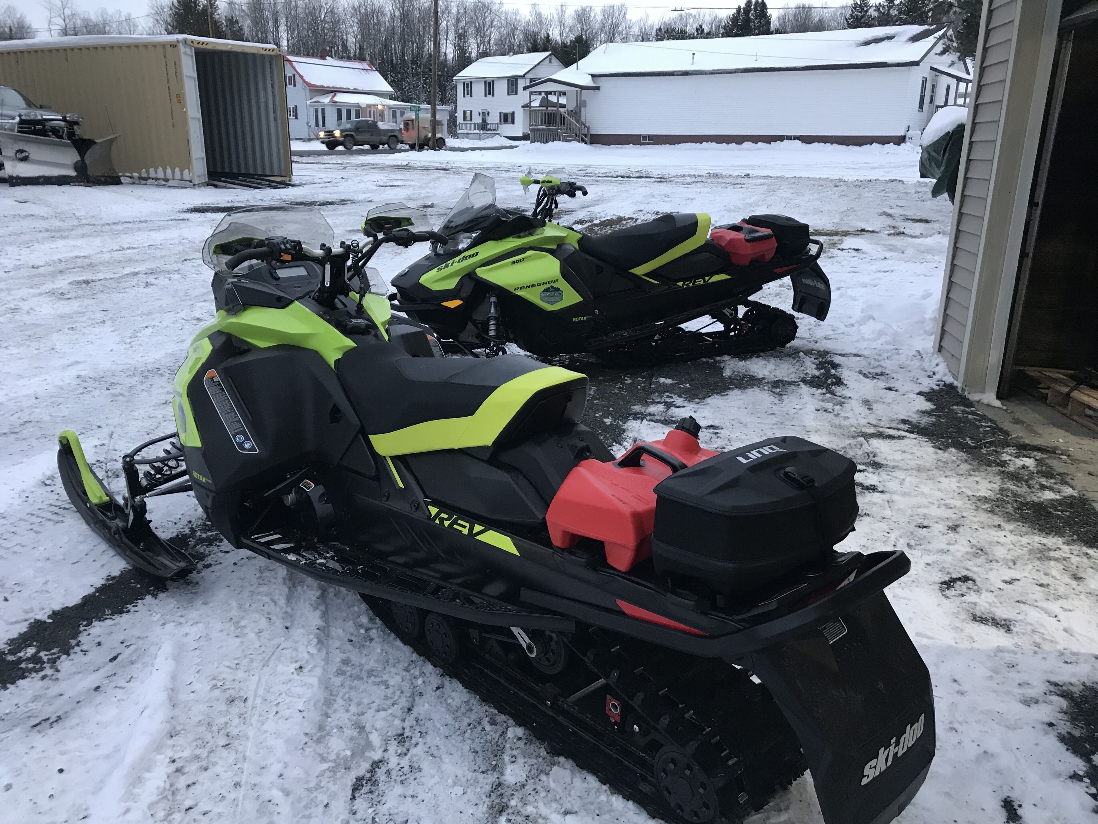 Two snowmobiles parked in a snowy area with a house and a garage in the background.