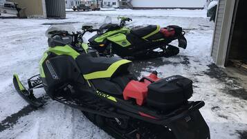 Two snowmobiles parked in a snowy area with a house and a garage in the background.