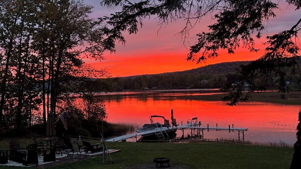 A lakeside view with a dock, chairs, and a table under a sunset sky. - Majestic Recreational Lodging w/ Snowmobile and Side x Side rentals on Long Lake (Sinclair)