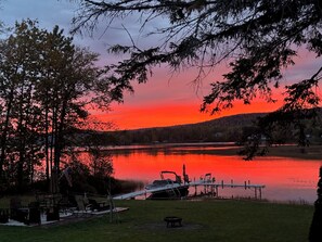 A lakeside view with a dock, chairs, and a table under a sunset sky. - Majestic Recreational Lodging w/ Snowmobile and Side x Side rentals on Long Lake (Sinclair)