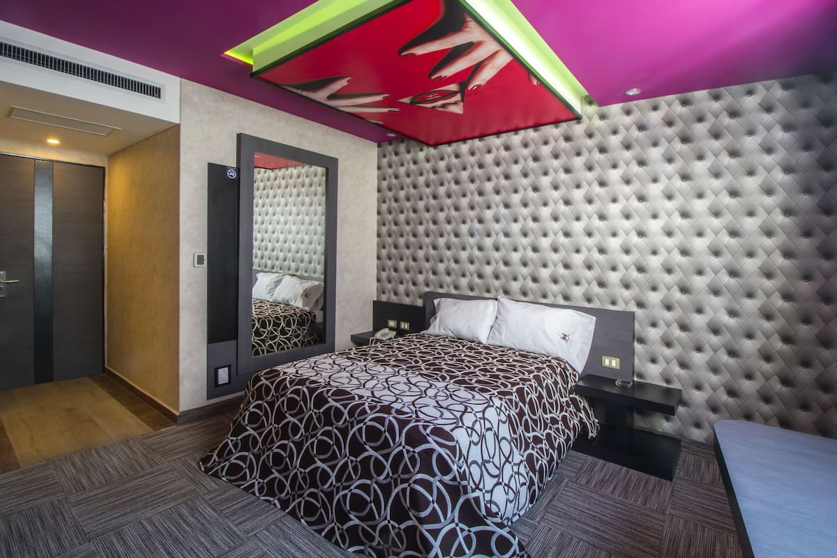 A modern hotel room with a tufted headboard, a patterned bedspread, a nightstand with a lamp, and a large mirror.