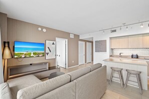 A modern living room with a large flat-screen TV, a sofa, and a kitchen area with a bar. - Panoramic bay views meet modern elegance in this 1-bedroom retreat (Hollywood)