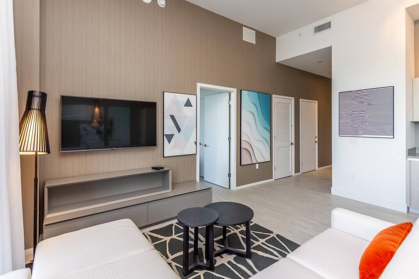 Un salon moderne avec un téléviseur à écran plat, un canapé, une table basse et une lampe. Les murs sont ornés d’œuvres d’art abstraites et une porte donne accès à une autre pièce.