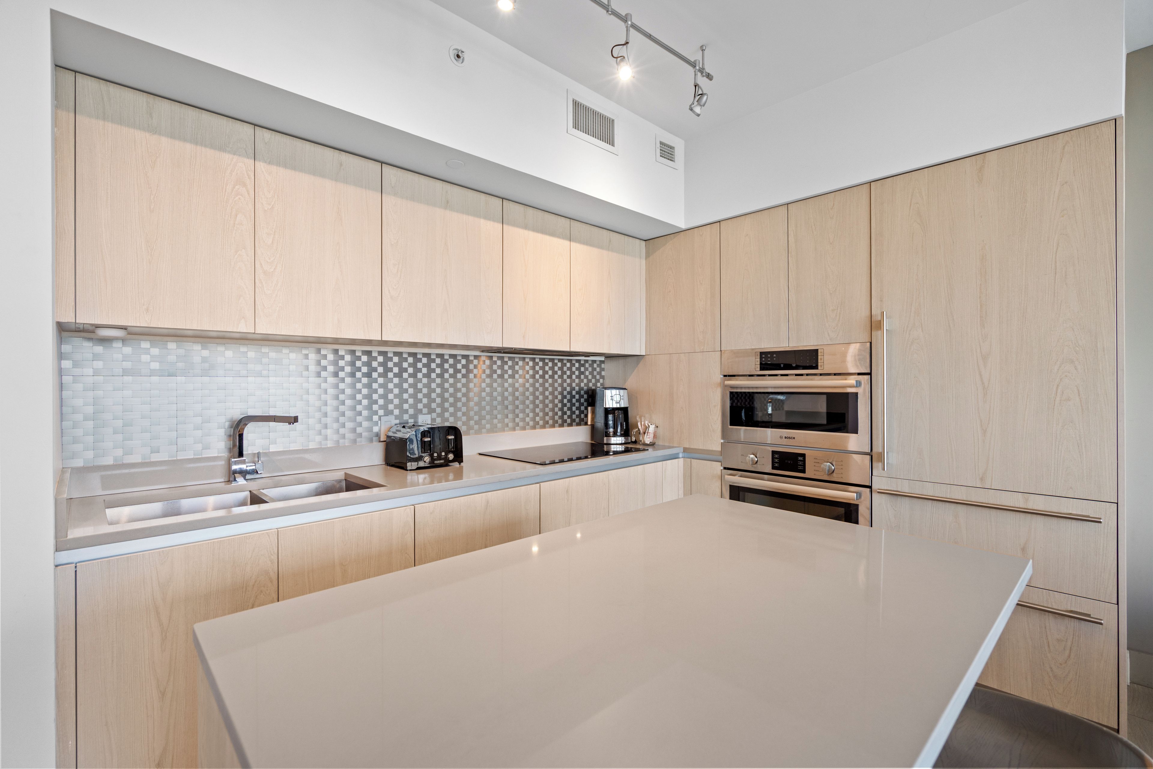 A modern kitchen with light wood cabinets, a large island, stainless steel appliances, and a tiled backsplash.