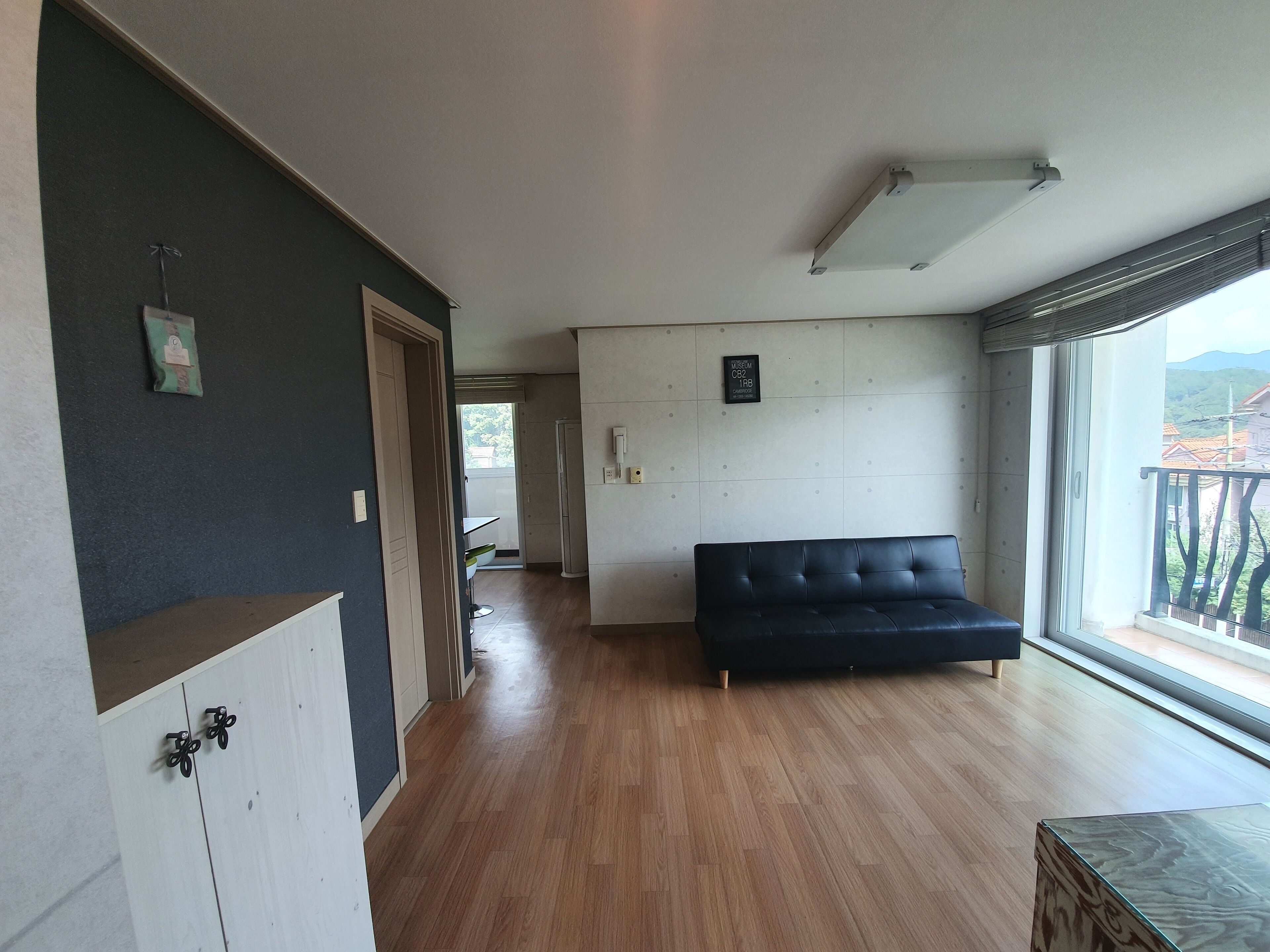 A modern living room with a dark sofa, wooden flooring, and a large window offering a view of the outdoors.