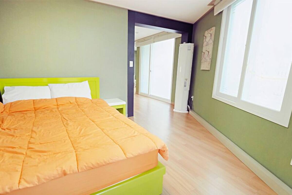 Une chambre à coucher avec un lit, une table de chevet verte, une armoire blanche et une grande fenêtre.