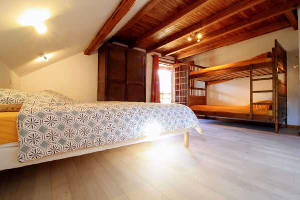 Une chambre avec un lit superposé, un grand lit avec une literie à motifs, des poutres en bois au plafond et une fenêtre avec des rideaux.