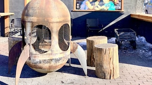 A large, rustic outdoor sculpture with a spherical body and two circular openings, surrounded by wooden stumps and a metal cart, set against a building with a mural.