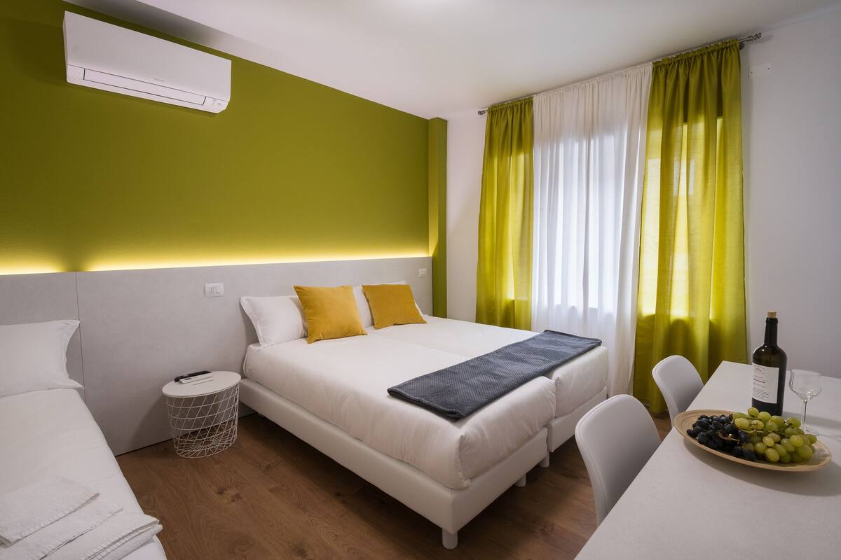 A modern bedroom with a bed, bedside tables, a small round table with a bottle and glasses, and a window with curtains.
