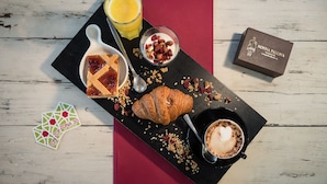 A breakfast tray with a croissant, coffee, a glass of orange juice, a bowl of yogurt with berries, and a chocolate box on a wooden table. - Zona 30 Bike Guesthouse (Palmanova)