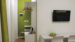 A room with a green accent wall, a flat-screen TV mounted on the wall, a small table with a bowl of apples, and a chair. - Zona 30 Bike Guesthouse (Palmanova)