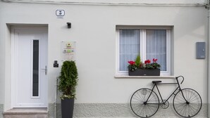 A white building with a door, a window with a basket of flowers, and a bicycle parked outside. - Zona 30 Bike Guesthouse (Palmanova)