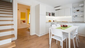 A modern kitchen with white cabinetry, a dining table with chairs, and a staircase with wooden steps. - Zona 30 Bike Guesthouse (Palmanova)