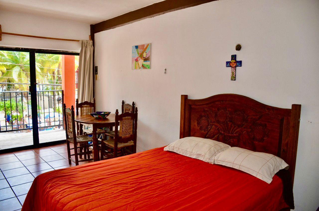A bedroom with a large bed, a wooden headboard, a small table with chairs, and a window with a view of palm trees.
