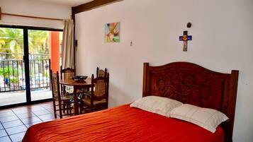 A bedroom with a large bed, a wooden headboard, a small table with chairs, and a window with a view of palm trees.