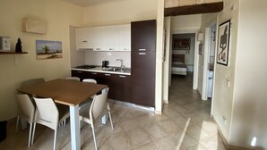 A compact kitchen with white cabinets, a dining area with a wooden table and chairs, and a hallway leading to a bedroom.