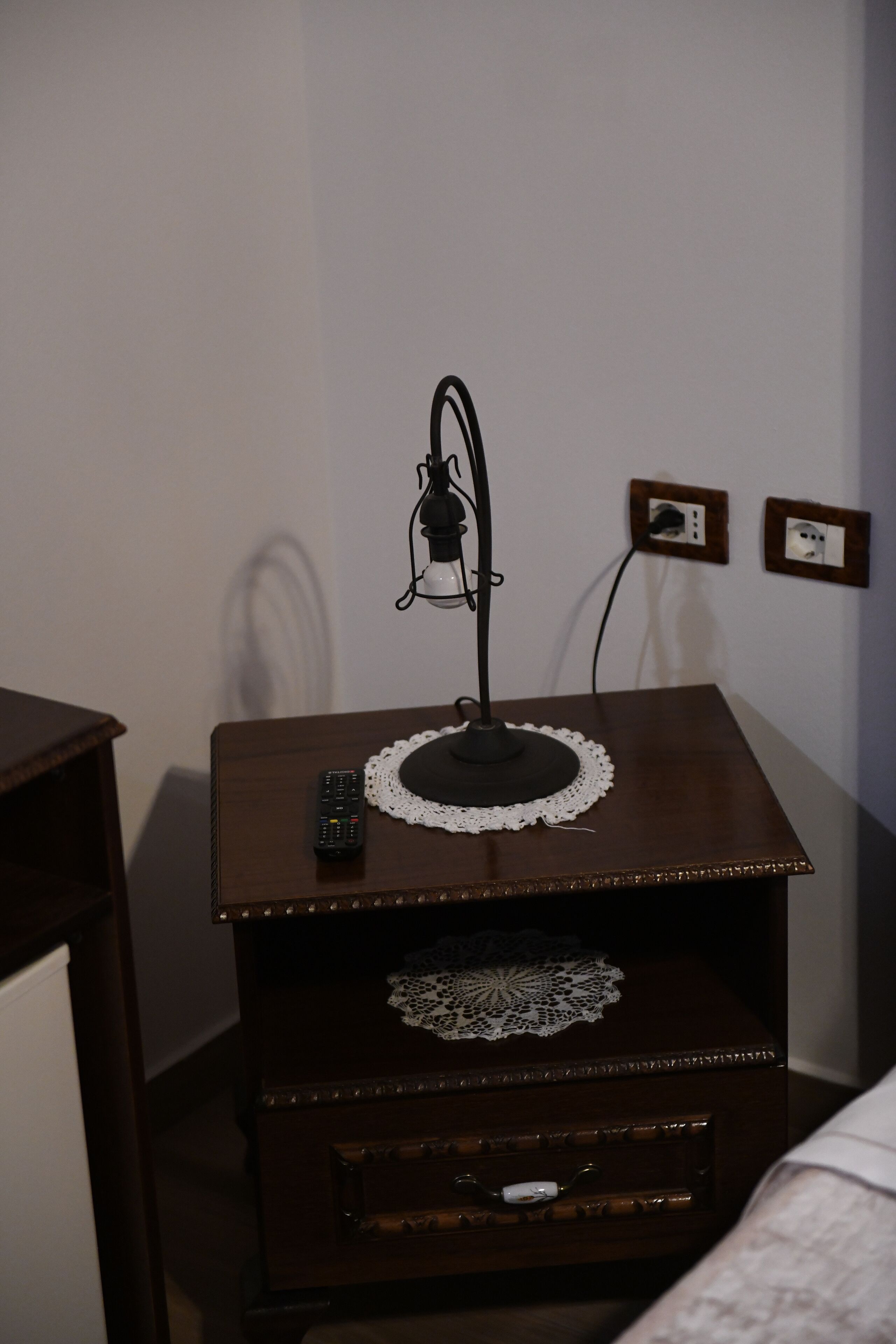 A wooden nightstand with a remote control, a lamp, and a power outlet on the wall.
