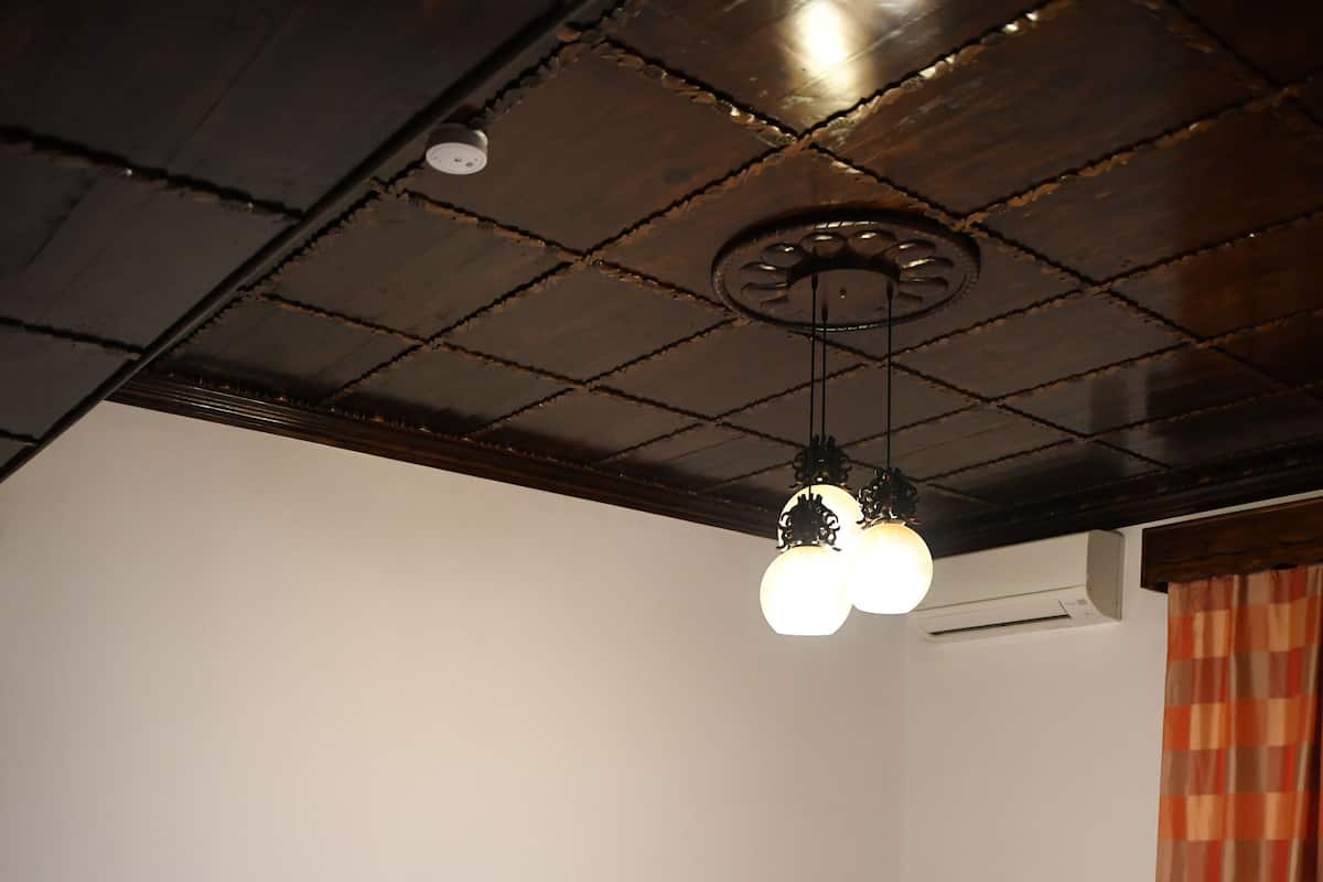A room with two wooden beds, each with a floral bedspread and matching pillows. There is a ceiling light fixture above the beds, and an air conditioning unit mounted on the wall.