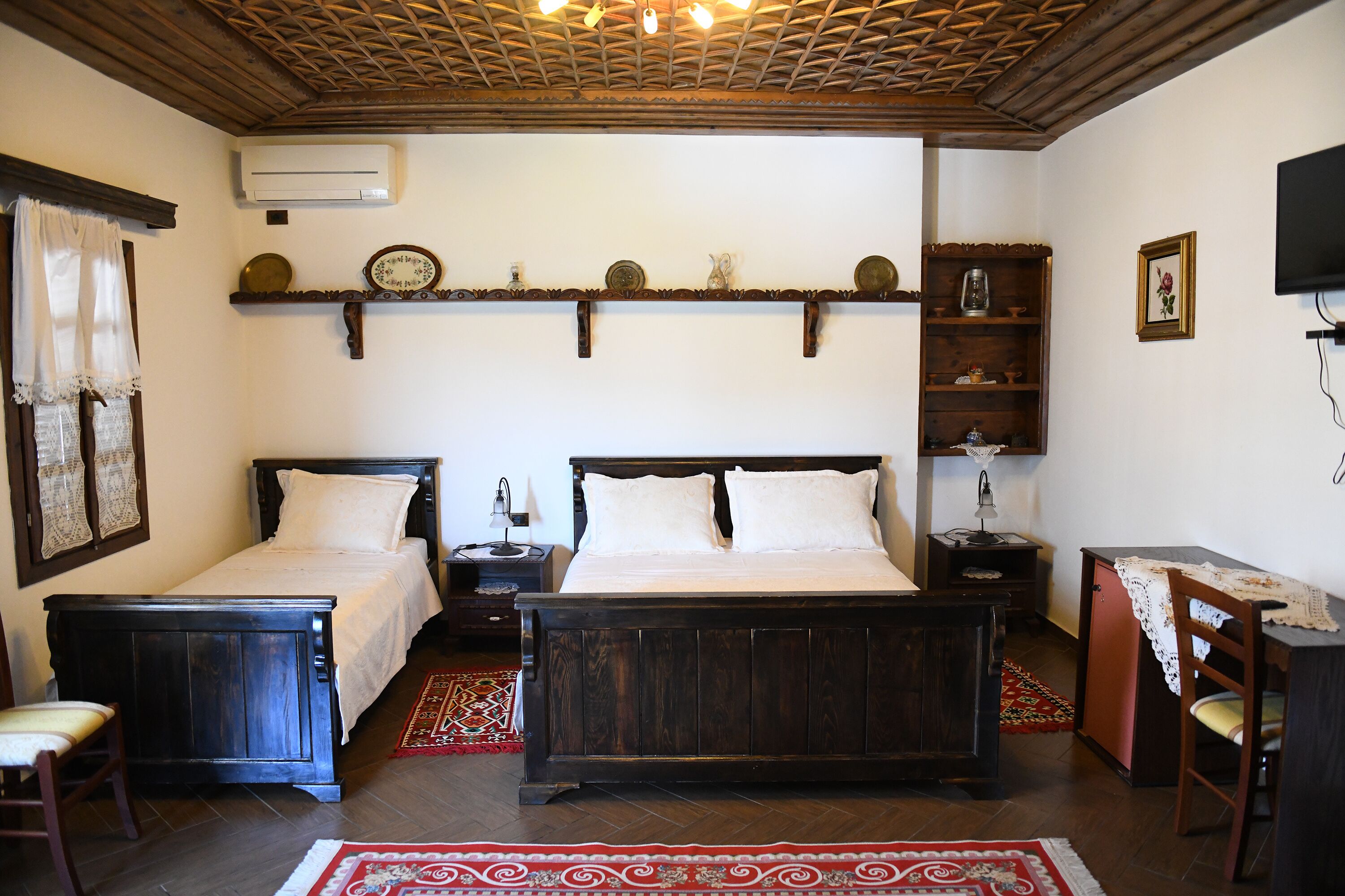 Une chambre avec deux lits, un plafond en bois, une étagère ornée d’assiettes décoratives, un téléviseur et une petite table avec une chaise.
