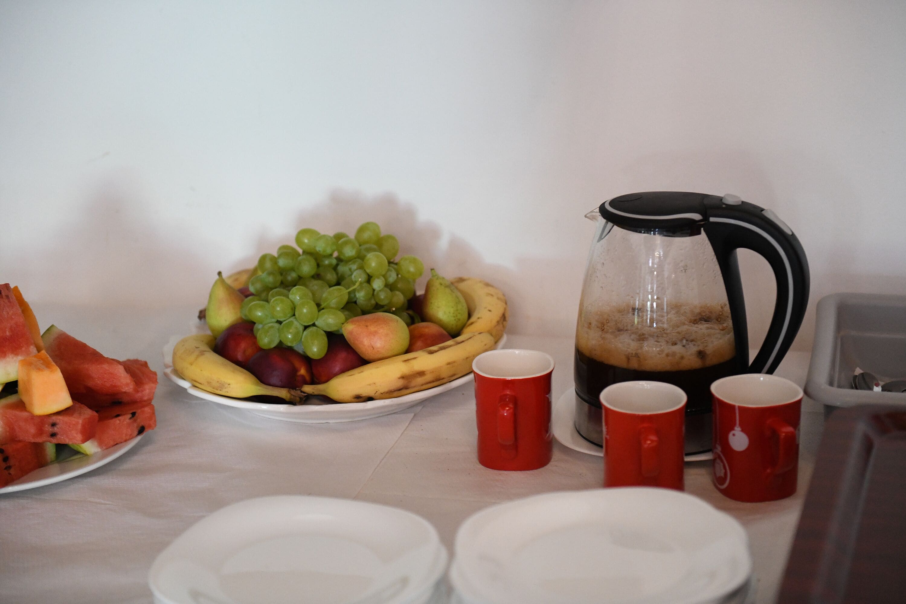 Une table avec un plateau de fruits, une cafetière et des tasses rouges.