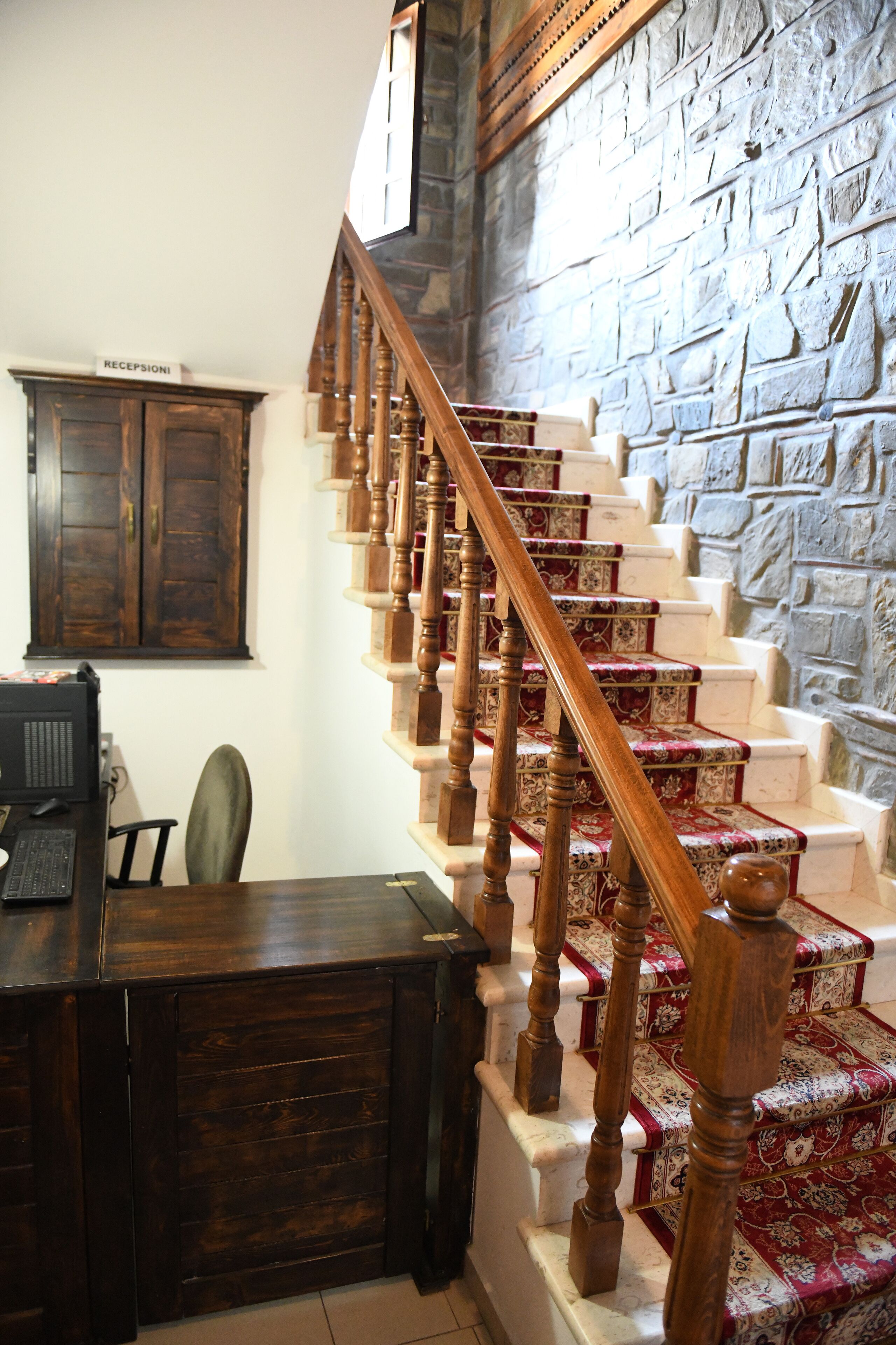Un escalier avec des balustrades en bois et des marches recouvertes d’une moquette rouge, menant à un palier où se trouvent un comptoir d’accueil et un meuble en bois.