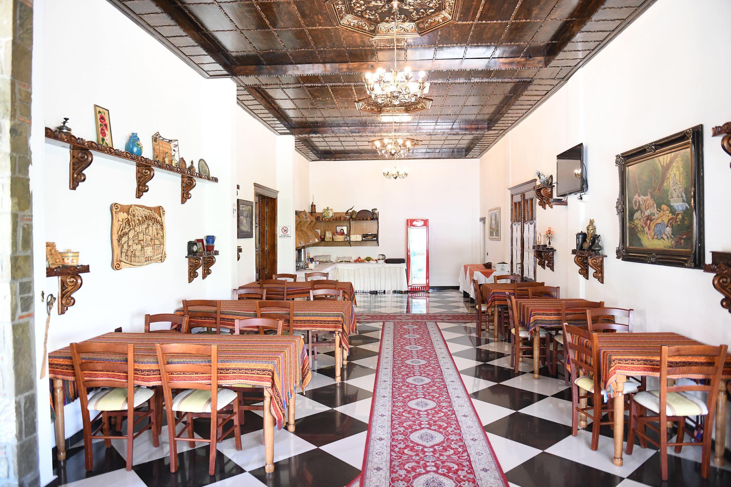 A dining hall with a checkered floor, wooden tables, and chairs, a chandelier, and wall-mounted decorations.
