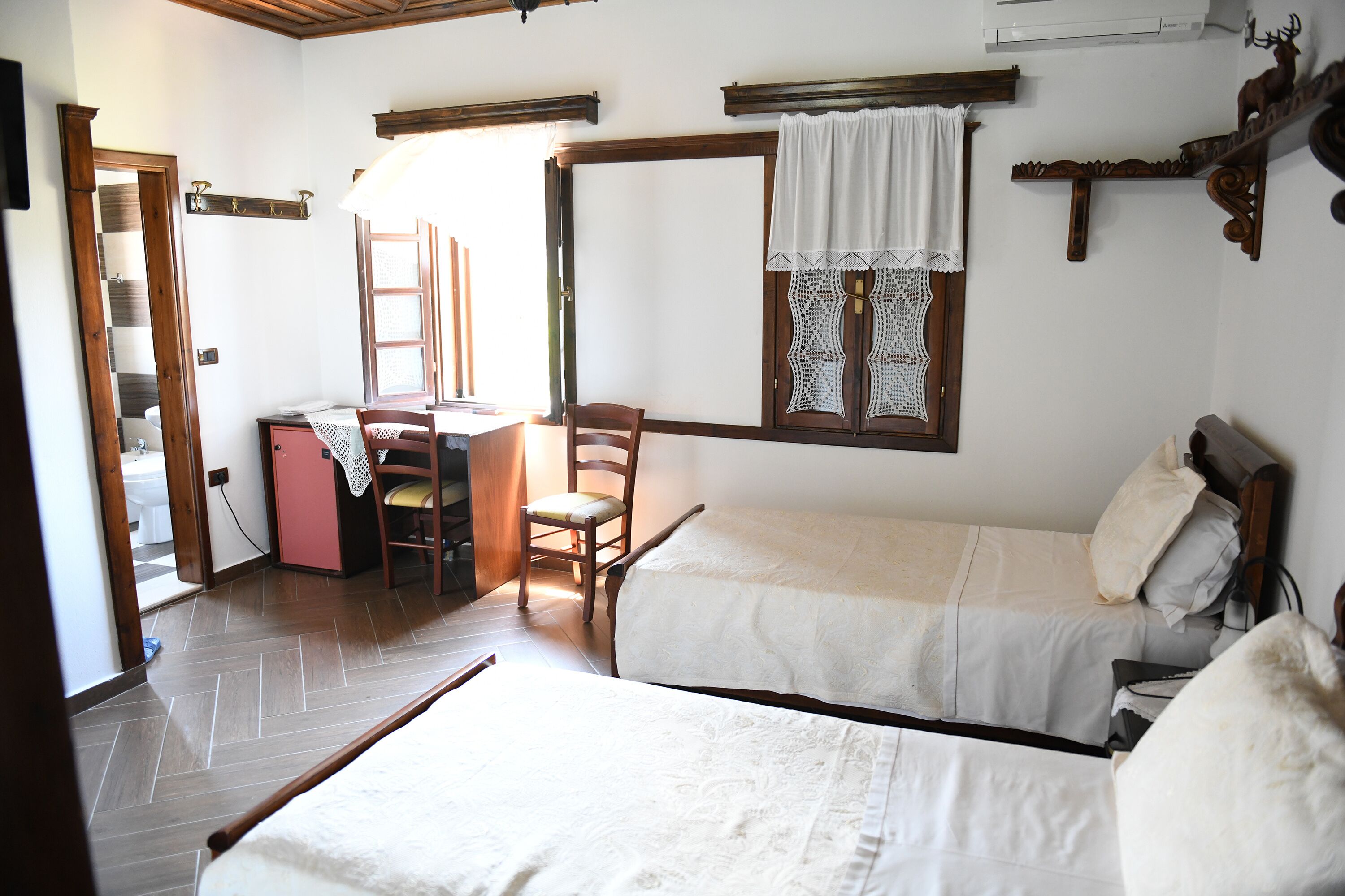 A room with two beds, a desk, and a chair. There is a window with a curtain, a mirror, and a shelf with a deer head decoration.