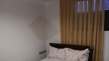 A bedroom with a wooden bed, a patterned bedspread, a hanging light fixture, a window with curtains, and a small bedside table.