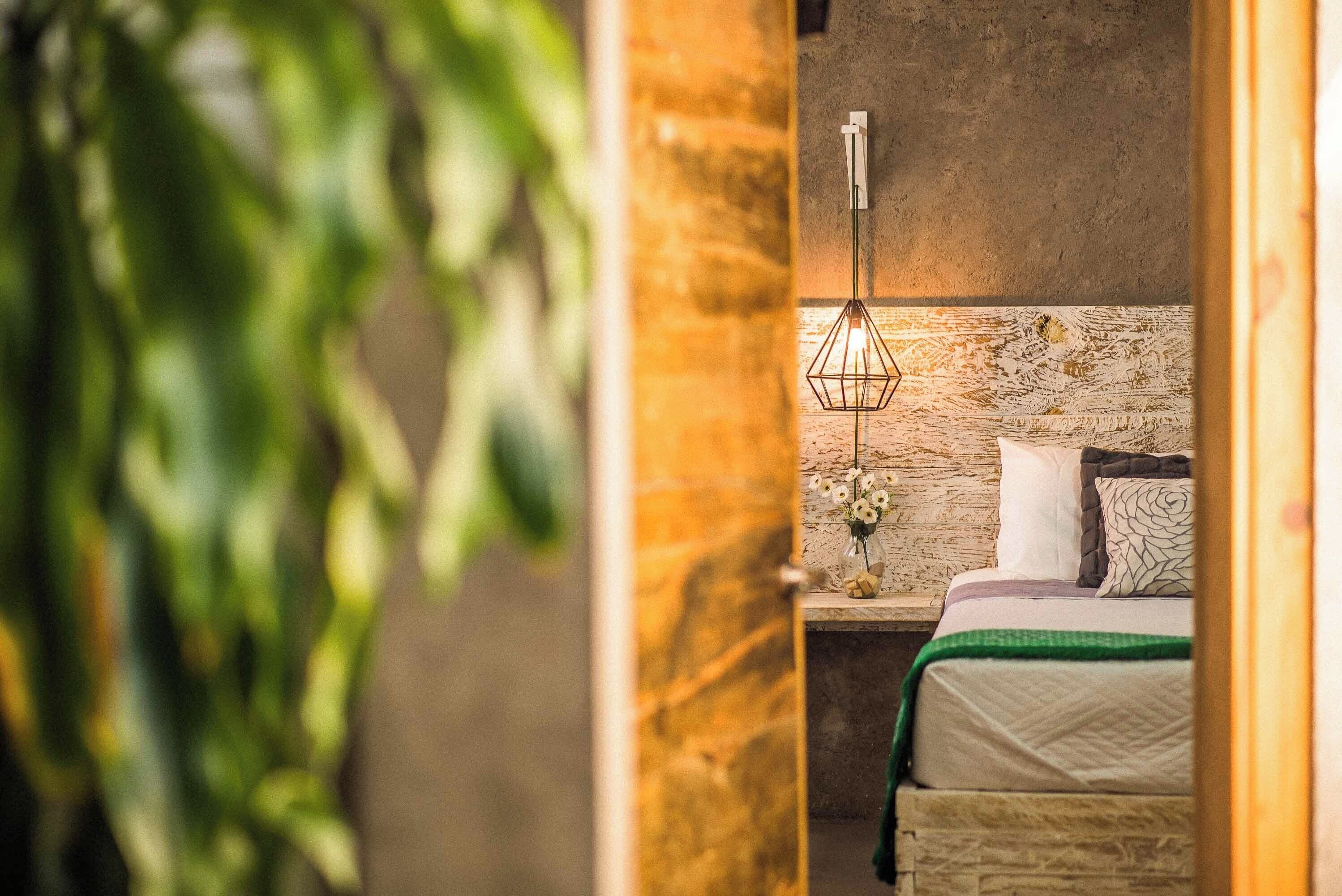 A bedroom with a stone headboard, a hanging light fixture, a bedside table with a vase of flowers, and a bed with a green throw.