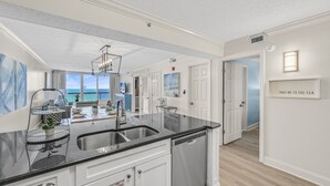 A modern kitchen with a black countertop, stainless steel sink, and white cabinetry. There is a view of the ocean through large windows. - ⛱️Pelican Beach 1915 - Cloud Nine | Gulf Views | 3 Pools⛱️ (Destin)