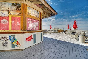 Uma cabana de praia de madeira com um anúncio de Coca-Cola e Budweiser, um mural com tema de prancha de surf e uma placa de boas-vindas para o Pelican Beach Tiki Hut.