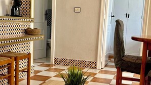 A room with a checkered floor, a wooden table with a plant, a chair, a shelf with a basket, a framed picture on the wall, and a door with a towel hanging on it.