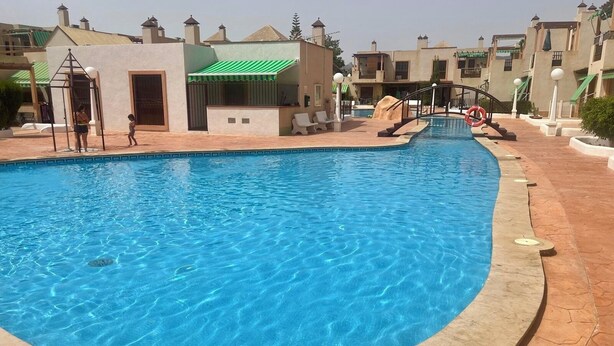 A swimming pool with a curved edge, surrounded by a tiled deck. There are buildings with balconies and a lifeguard stand in the background.