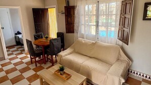 A living room with a sofa, a dining table with chairs, a ceiling fan, and a window with curtains.
