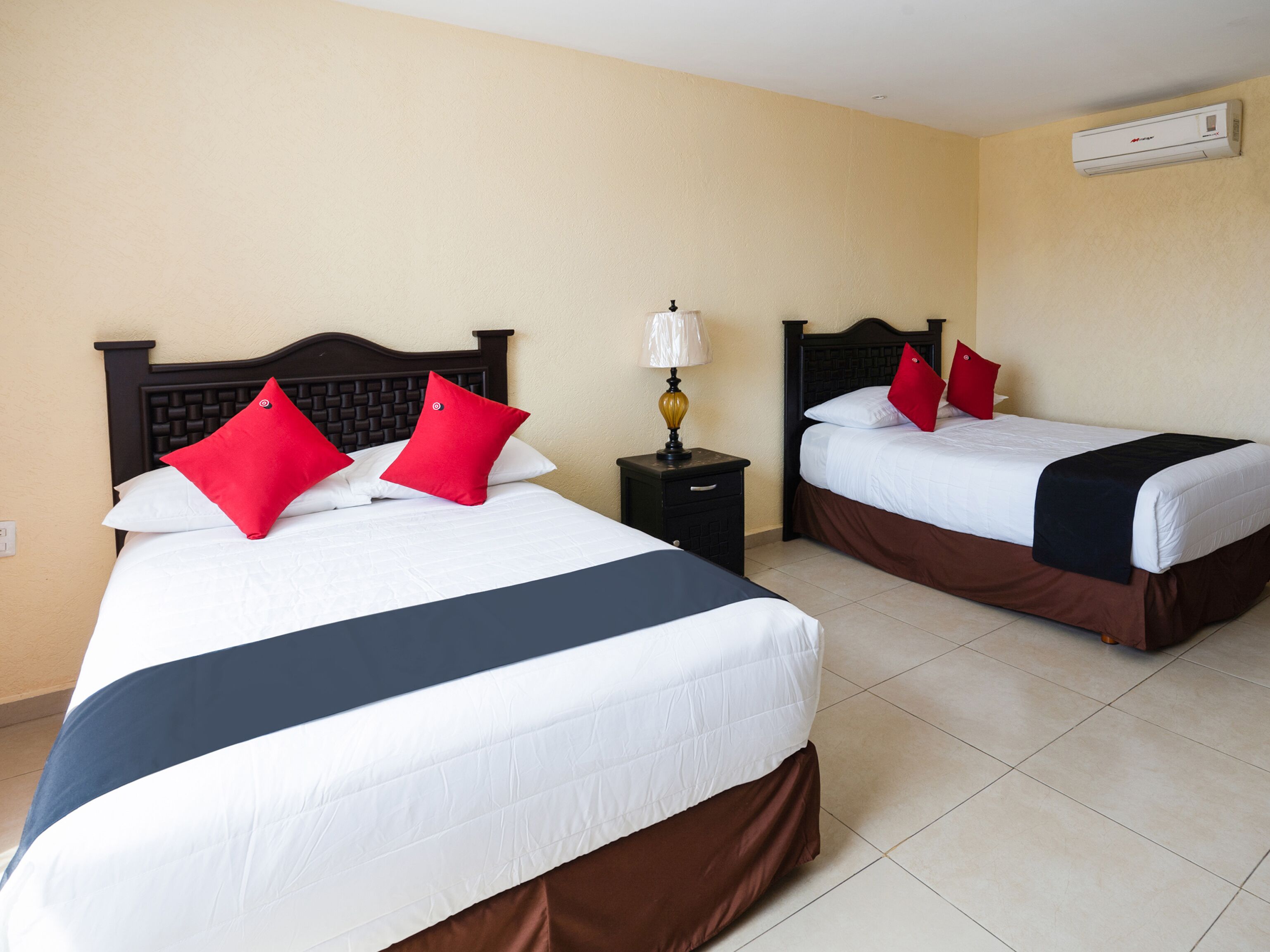 Two double beds with red and white bedding, a nightstand with a lamp, and an air conditioning unit on the wall.