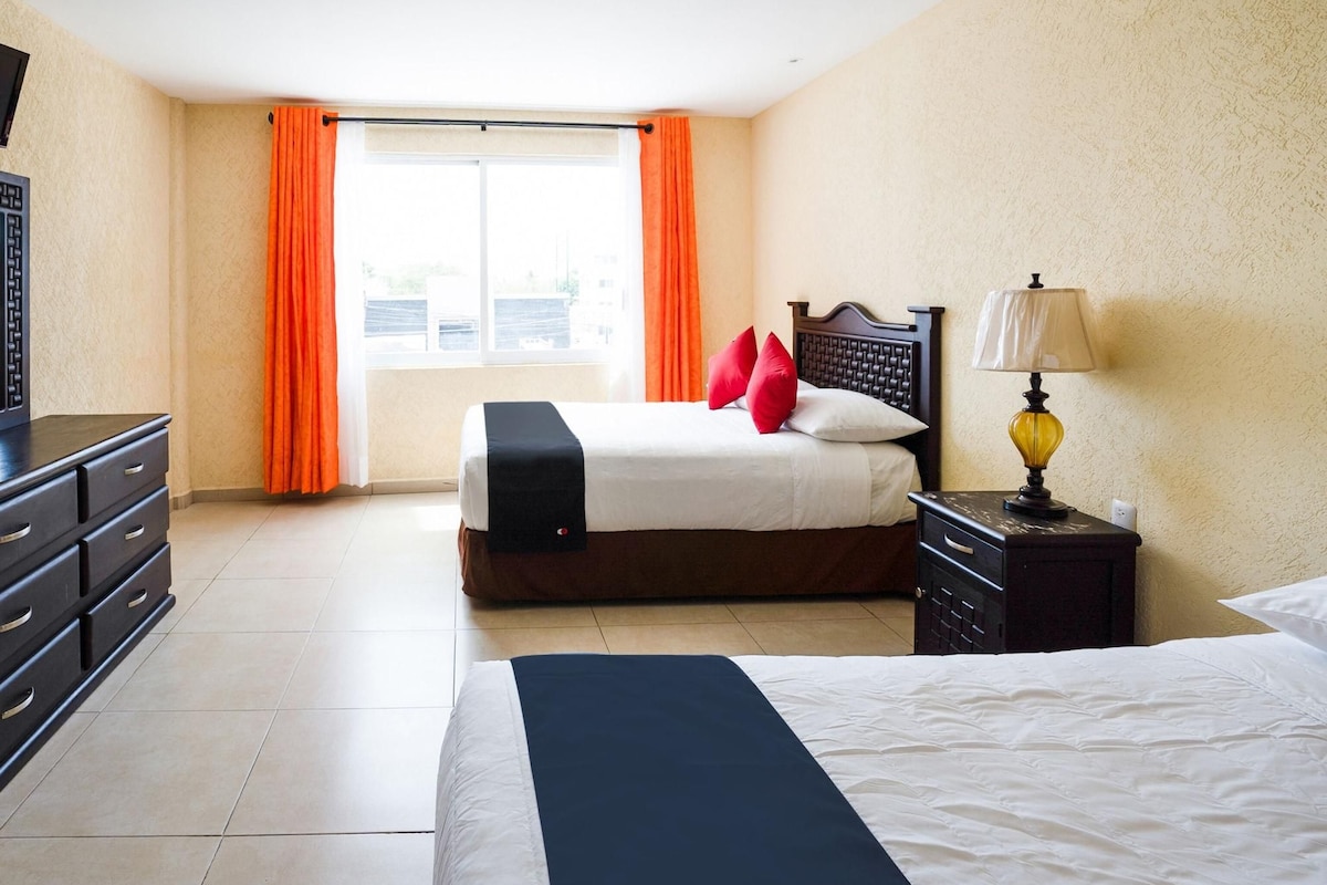 A hotel room with a bed, a nightstand with a lamp, a dresser, and a window with curtains.