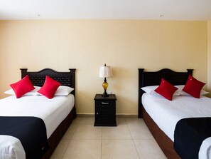 Ein Hotelzimmer mit zwei Betten, jeweils mit schwarzem Kopfteil, weißer Bettwäsche und roten Kissen. Dazwischen befindet sich ein Nachttisch mit einer Lampe.