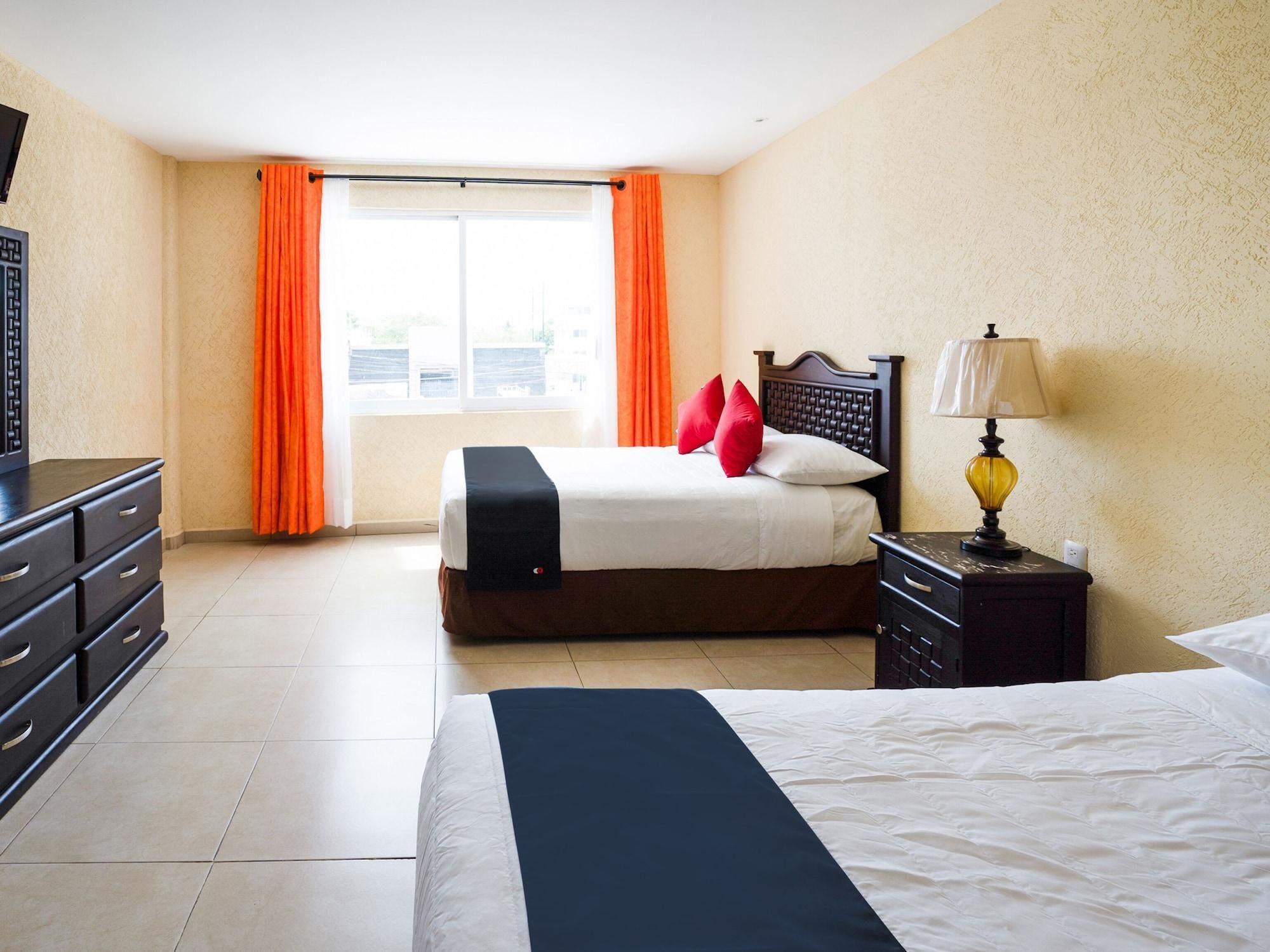 A hotel room with a bed, a nightstand with a lamp, a dresser, and a window with curtains.
