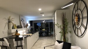 A modern kitchen with a central island, bar stools, a dining area with a black chair and white bowl, and a large wall clock. - Private Vacation Home w/ Heated Pool. Walk/Bike to Vanderbilt Beach & Amenities. (Naples)