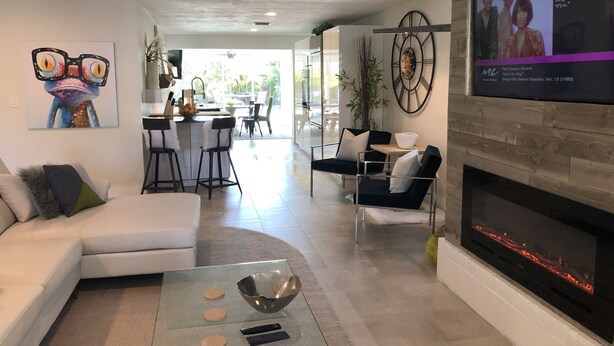 A modern living room with a white sofa, a glass coffee table, and a fireplace. There is a kitchen area in the background with a dining table and chairs. - Private Vacation Home w/ Heated Pool. Walk/Bike to Vanderbilt Beach & Amenities. (Naples)