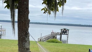 Eine Hängematte, die an einem Baum befestigt ist, ein Picknicktisch mit einer Weinflasche und Gläsern sowie ein Blick auf einen See mit einem Steg und einer Mole im Hintergrund.