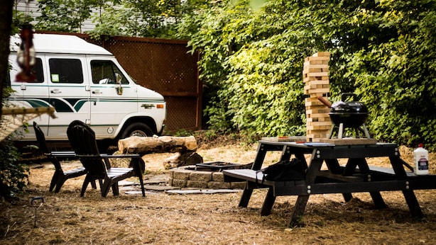 A rustic outdoor setting with a white van, picnic table, and chairs. - Van Glamping - AC, Heat and Power! (Landrum)
