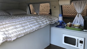 A bedroom with a bed, a nightstand with a microwave, and string lights on the ceiling. - Van Glamping - AC, Heat and Power! (Landrum)