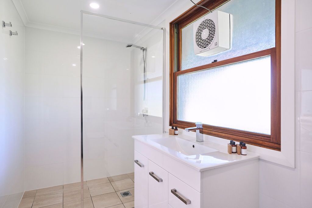Un baño moderno con cabina de ducha de vidrio, tocador blanco con lavabo y una ventana sobre el espejo.