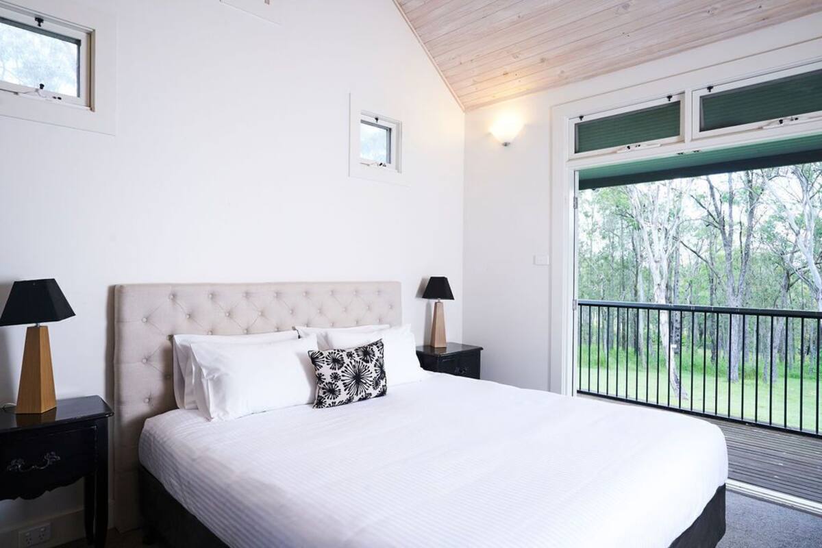 A bedroom with a large bed, two bedside tables, a window, and a balcony with a view of trees.