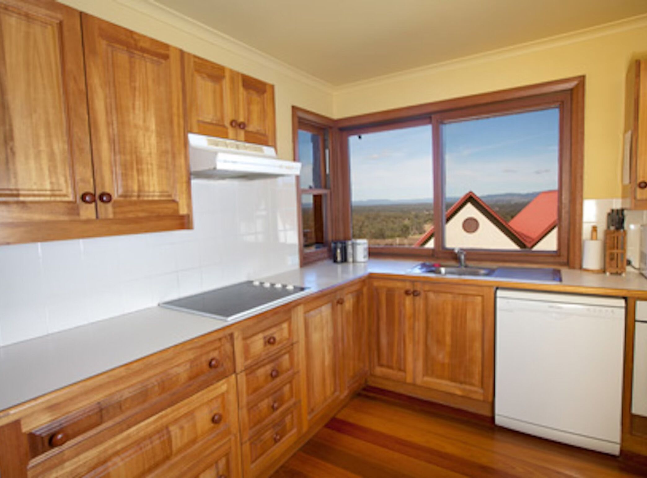 A kitchen with wooden cabinets, a white countertop, a dishwasher, and a window with a view of a house and landscape.